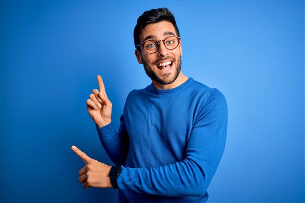 مرد جوان خوشتیپ با ریش ژاکت و عینک معمولی روی پس‌زمینه آبی لبخند می‌زند و به دوربین نگاه می‌کند که با دو دست و انگشت به پهلو اشاره می‌کند