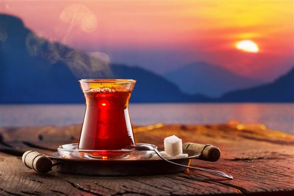 لیوان با چای سنتی ترکی سیاه روی یک میز چوبی قدیمی ایستاده است پشت غروب خورشید در سواحل دریای ترکیه