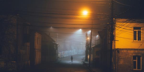 زن تنها در حال قدم زدن در شهر قدیمی مه آلود با چراغ های خیابان در کت