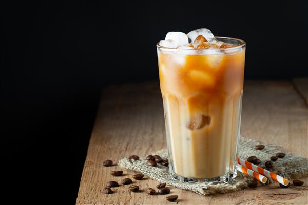 قهوه یخی در یک لیوان بلند با خامه ریخته شده روی آن تکه های یخ و دانه ها روی یک میز چوبی قدیمی روستایی نوشیدنی سرد تابستانی با لوله در پس زمینه مشکی با فضای کپی