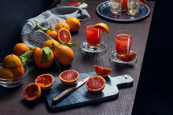 آب پرتقال خونی سیسیلی تازه فشرده شده در لیوان هایی با پرتقال برش خورده روی میز چوبی