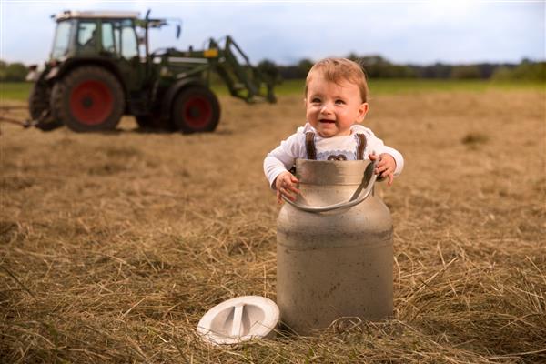 یک نوزاد پسر باواریایی ستایش‌انگیز که در یک شیرخوار ایستاده و با خوشحالی می‌خندد در پس‌زمینه تراکتوری در حال چرخاندن یونجه‌ای است که روی آن شیر می‌ریزد