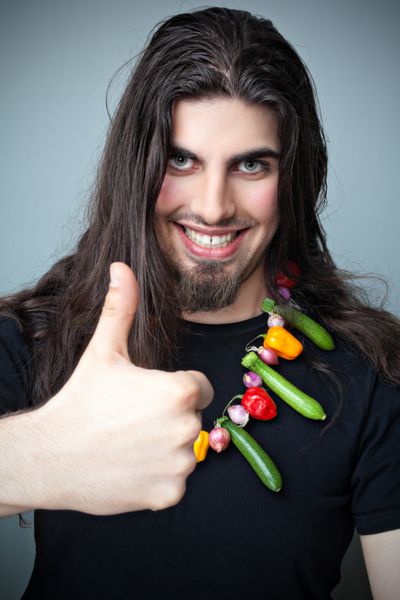 مرد جوان قفقازی جذاب با موهای بلند تیره که گردنبندی از سبزیجات مینیاتوری به تن دارد و انگشت شست به بالا