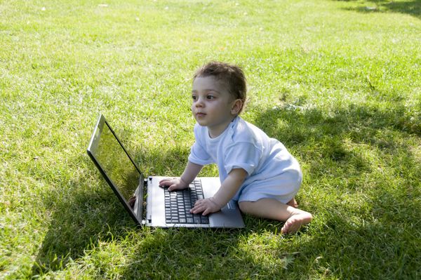 کودک شاد که با لپ تاپ روی چمن ها سرگرم می شود