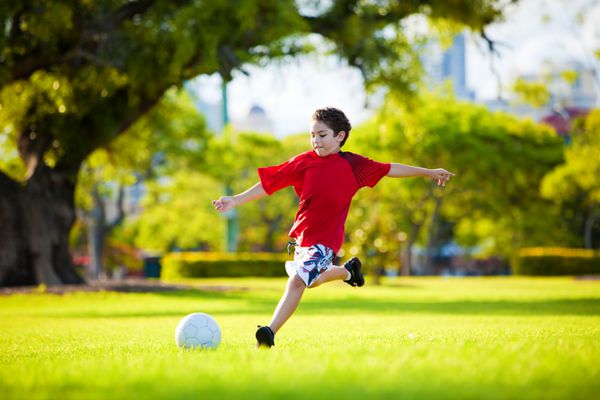 پسر جوان هیجان زده در حال لگد زدن به توپ در چمن در فضای باز