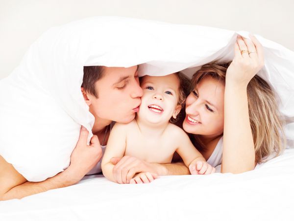 خانواده شاد مادر پدر و نوزادشان زیر پتو روی تخت در خانه تمرکز روی مرد