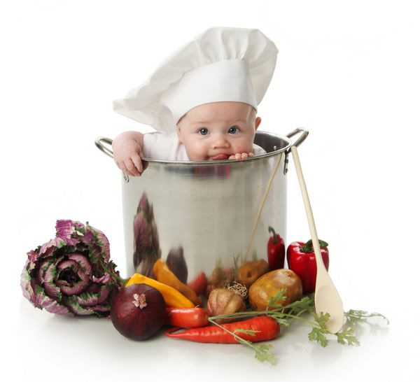 پرتره کودکی نشسته با کلاه سرآشپزی که داخل آن نشسته است و یک قابلمه بزرگ آشپزی احاطه شده با سبزیجات و غذا را می لیسد جدا شده روی سفید