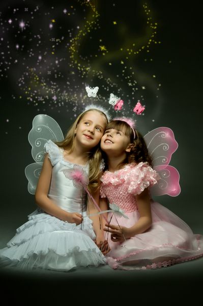 دو دختر کوچک زیبا با بال بنشینید و روی پس زمینه تیره لبخند بزنید