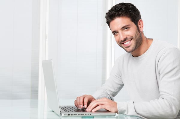 مرد جوان راضی خوشحال که روی لپ تاپ کار می کند و به دوربین نگاه می کند