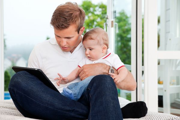 پدری که کودک را در آغوش می گیرد در حالی که از تبلت دیجیتال در خانه استفاده می کند