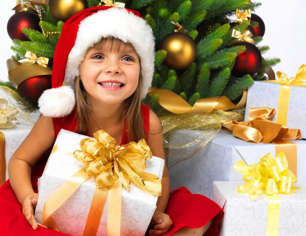 دختر کوچک زیبا با هدیه نزدیک درخت کریسمس لبخند می زند