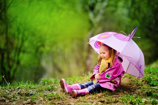 دختر کوچک خندان با چتر با کت بارانی و چکمه در فضای باز