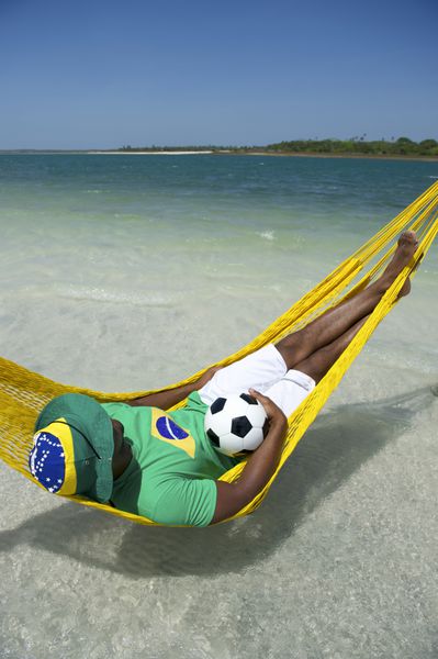 مرد برزیلی در حال استراحت در بانوج ساحلی با توپ فوتبال خود و لباس پرچم برزیل