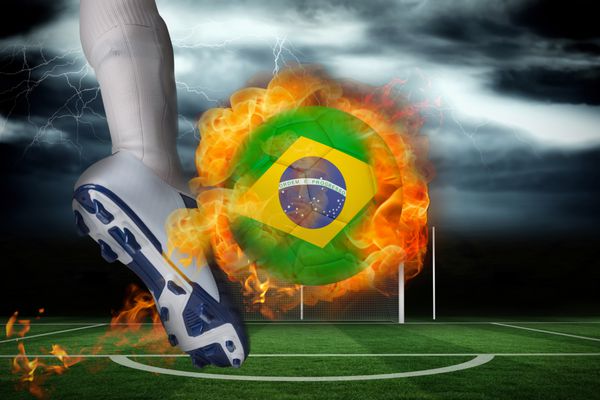 بازیکن فوتبال در حال لگد زدن به توپ شعله ور پرچم برزیل در مقابل زمین فوتبال در زیر آسمان طوفانی