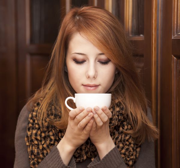 دختر مو قرمز سبک در حال نوشیدن قهوه در نزدیکی درهای چوبی