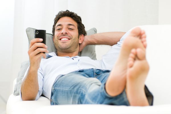 مرد جوان خندان در حال گشت و گذار در اینترنت و ارسال پیامک با تلفن همراه در خانه