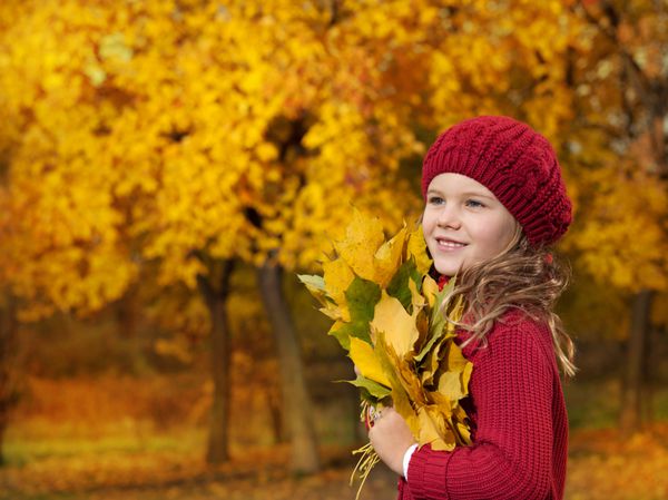 پرتره دختر جوان قفقازی جذاب با لباس های رنگارنگ گرم روی برگ های زرد در فضای باز در حال خنداندن به بالا