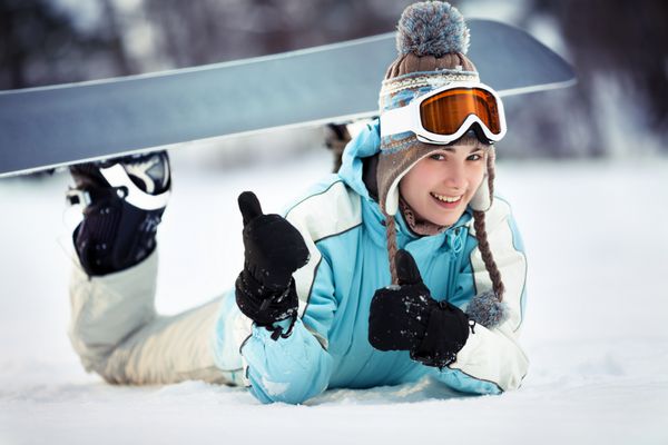 اسنوبورد زن جوان زیبا در حال استراحت در پیست اسکی در جلو دراز کشیده است شست را نشان می دهد و لبخند می زند از نزدیک