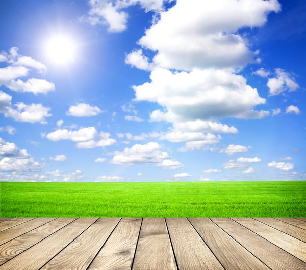زمین سبز تابستانی زیبا با آسمان آبی با ابرهای خاکستری و خورشید درخشان و تخته های چوبی روی زمین