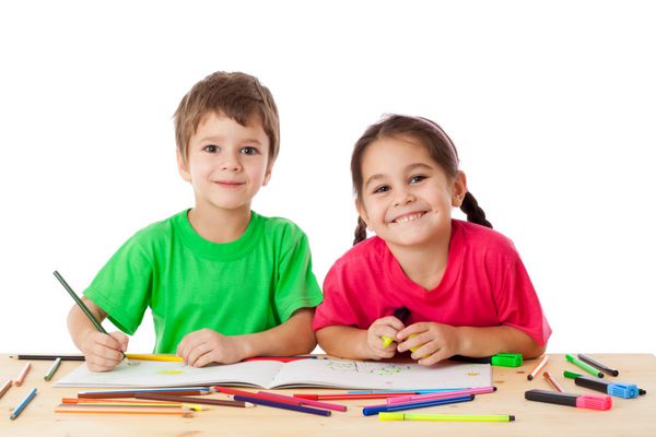 دو بچه کوچک خندان پشت میز با مداد رنگی جدا شده روی سفید نقاشی می‌کنند