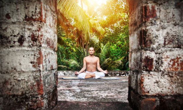 مدیتیشن یوگا توسط مردی با ریش با شلوار سفید روی پشت بام در کف دست و پس‌زمینه آسمان غروب خورشید در هند