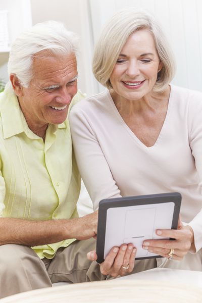 زن و شوهر سالخورده خوشبختی که با هم در خانه روی مبل با استفاده از رایانه لوحی نشسته اند