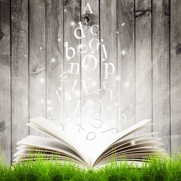 کتاب باز با حروف پرنده در چمن سبز روی پس‌زمینه چوبی کتاب جادو