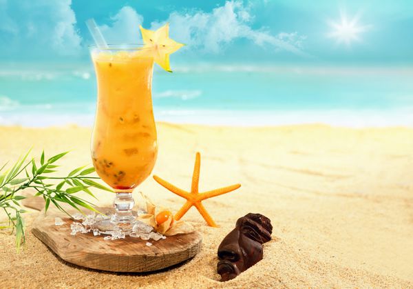 کارامولای رنگارنگ و کوکتل نارنجی در یک لیوان بلند که روی یک تخته چوبی در ساحل طلایی در یک استراحتگاه تعطیلات گرمسیری در طول تعطیلات تابستانی لذت بخش سرو می شود