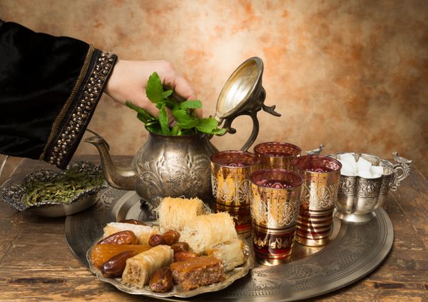 دست زن در حال افزودن برگ نعنا به سینی با خرما کلوچه و چای به سبک مراکشی