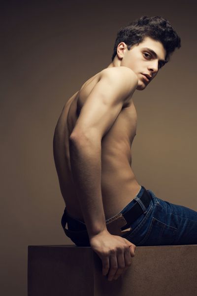 مدل مردانه عضلانی زیبا خوش تیپ با شلوار جین در حال ژست گرفتن و برهنه شدن پسری که روی یک مکعب چوبی نشسته است سبک Vogue فضای کپی پرتره استودیو مد