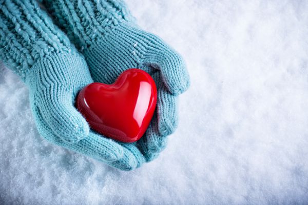 دست‌های زن با دستکش‌های بافتنی قهوه‌ای روشن قلب زیبای قرمز براق را روی پس‌زمینه برفی در دست گرفته است مفهوم عشق و سنت ولنتاین
