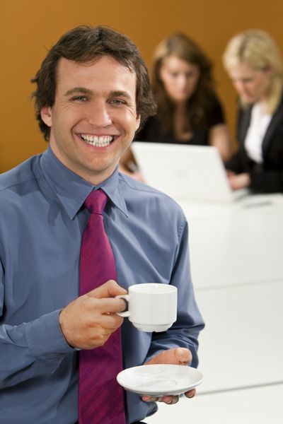 یک مدیر مرد جوان در حالی که همکارانش پشت سر او روی لپ‌تاپ کار می‌کنند یک قهوه استراحت می‌کند