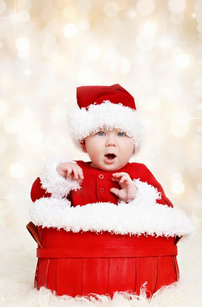 کودک شاد ناز با لباس‌های قرمز کریسمس بابانوئل که در یک سبد روی پس‌زمینه چراغ‌های بدون تمرکز نشسته است