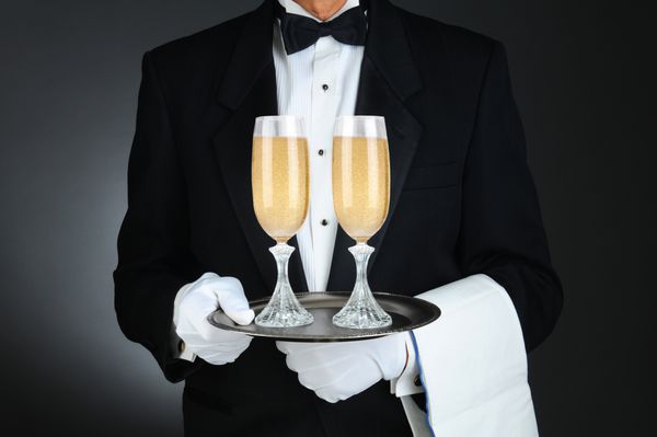 نمای نزدیک از یک سوملیه که دو لیوان شامپاین را روی سینی در جلوی تنه‌اش نگه داشته است فرمت افقی در زمینه خاکستری روشن تا تیره