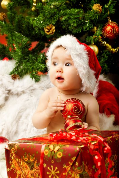 کودک کریسمس با کلاه که توپ قرمز را در نزدیکی جعبه هدیه و درخت صنوبر سال نو نگه داشته است