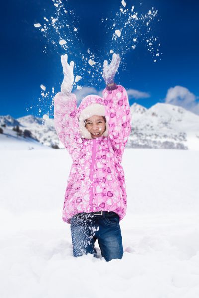 دختر کوچک خندان و شاد صورتی با برف در هوا پرتاب می کند و دانه های برفی در همه جهات پرواز می کنند
