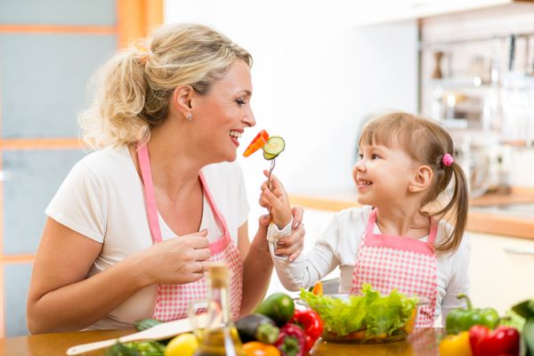 دختر بچه در حال تغذیه مادر با سبزیجات در آشپزخانه