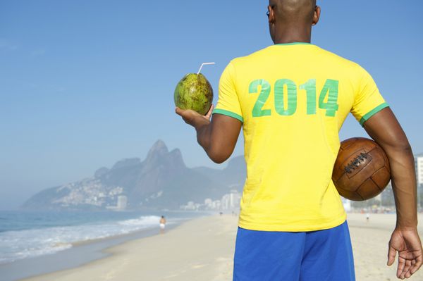 بازیکن فوتبال برزیلی با پیراهن سال 2014 به رنگ های برزیل در حالی که توپ فوتبال قدیمی را در دست دارد و در حال نوشیدن نارگیل ریودوژانیرو است