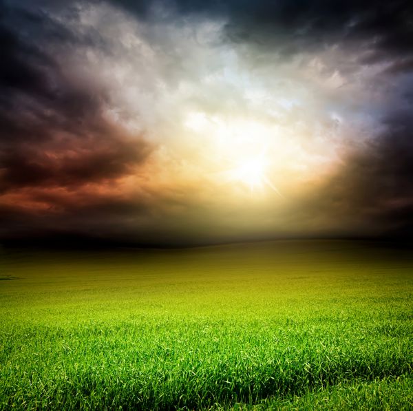 آسمان دراماتیک طوفانی و زمین سبز چمن با نور خورشید که در عصر از میان ابرها عبور می کند