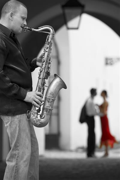 عکس سیاه و سفید یک نوازنده خیابانی در حال نواختن ساکسیفون خود در حالی که یک زوج رمانتیک خارج از فوکوس در پس زمینه دیده می شود که زن رنگی با لباس قرمز پوشیده است