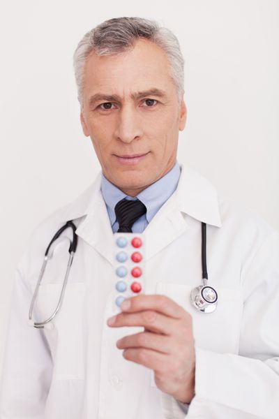 قرصاتو بخور دکتر ارشد موهای خاکستری با لباس فرم که قرص ها را در دست دارد و در حالی که روی سفید ایستاده است به دوربین نگاه می کند