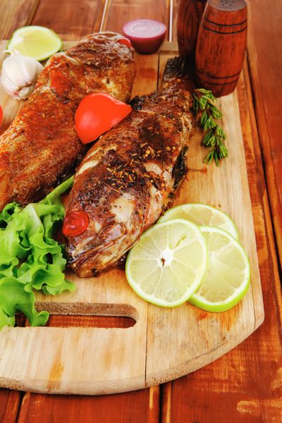 غذای سالم دو ماهی باس دریایی سرخ شده با گوجه فرنگی و سبزیجات روی تخته چوبی بزرگ روی میز سرو می شود