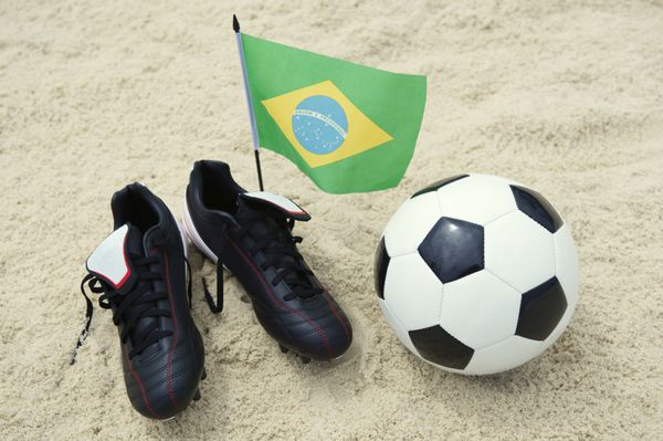 چکمه های فوتبال با توپ فوتبال و پرچم برزیل در پس زمینه ساحل شنی