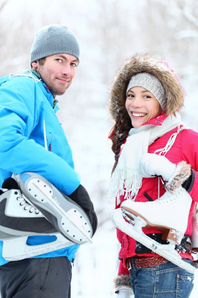 زوج زمستانی اسکیت روی یخ با خوشحالی در دست داشتن اسکیت روی یخ در فضای باز لبخند می زنند زوج جوان زیبا زن آسیایی مرد قفقازی در بیرون در روز زمستان برفی
