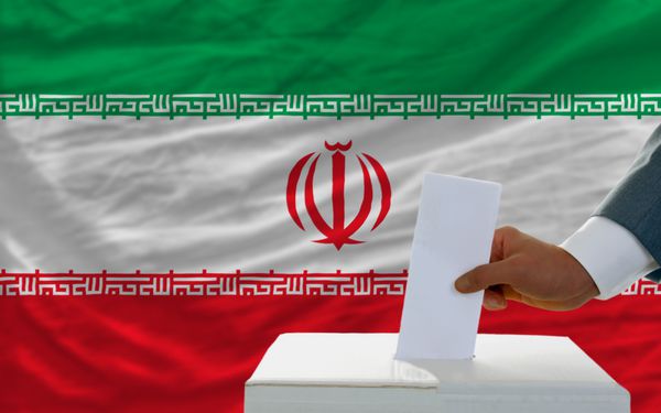 مردی در حال گذاشتن برگه رای در صندوق در هنگام انتخابات در ایران در مقابل پرچم
