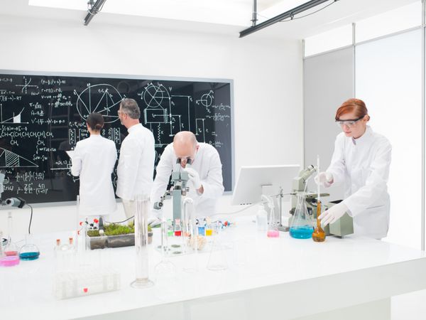 تیمی از دانشمندان در آزمایشگاهی که روی آزمایش‌های شیمیایی میکروسکوپ و بحث و تجزیه و تحلیل فرمول‌های مولکولی پیچیده روی تخته سیاه بزرگ کار می‌کنند
