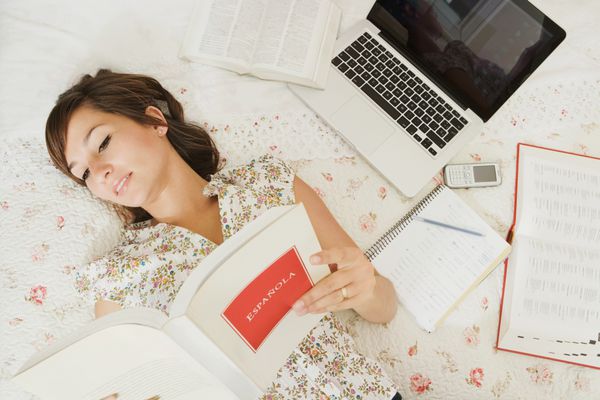 نمای بالای سر یک نوجوان دانش‌آموز زبان در حال انجام تکالیف روی تخت خوابش در اتاق خوابش با رایانه لپ‌تاپ و تلفن همراه در حال خواندن یک کتاب اسپانیایی باز