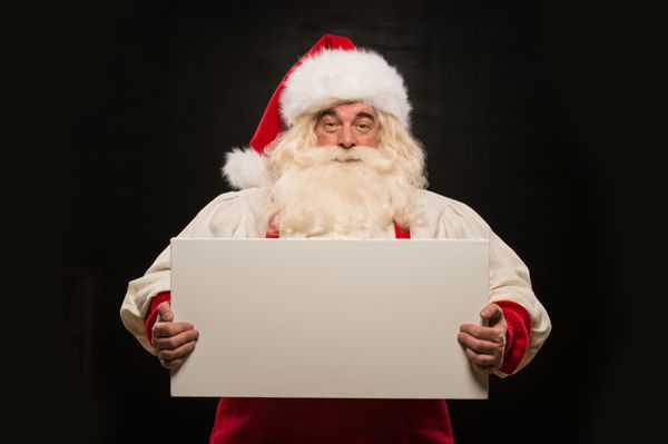 بابا نوئل با نگه داشتن تابلوی سفید سفید با سرگرمی و لبخند در مقابل پس زمینه تاریک ایستاده است