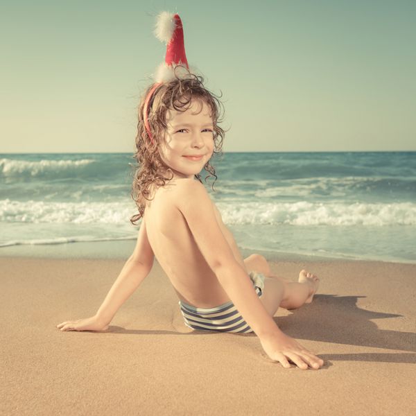 کودک شاد با کلاه بابانوئل در حال تفریح در ساحل استوایی مفهوم تعطیلات کریسمس