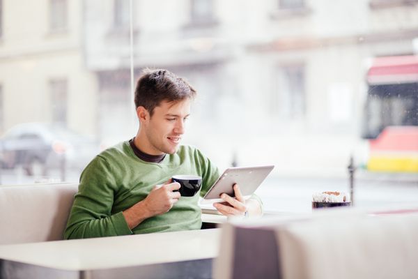 مرد جوان در حال نوشیدن قهوه در کافه و استفاده از رایانه لوحی